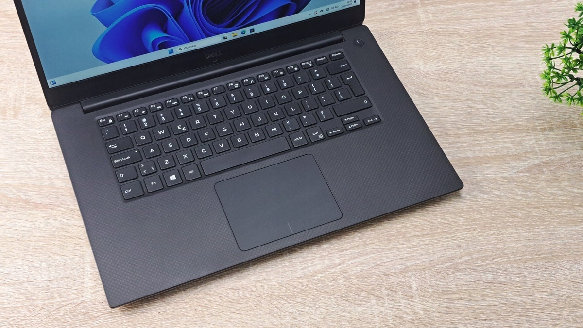 Zbliżenie z góry na otwartą klawiaturę i touchpad laptopa Dell w czarnej obudowie, ustawionego na blacie.