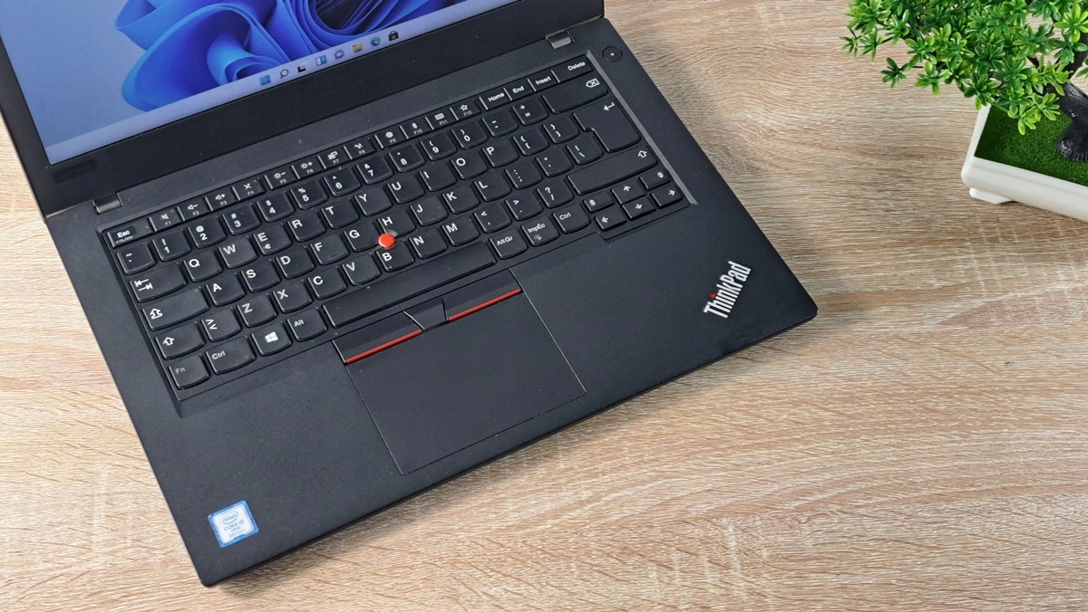 Otwarty laptop ThinkPad z widoczną klawiaturą, czerwonym trackpointem i touchpadem, ujęcie z góry na drewnianym biurku.