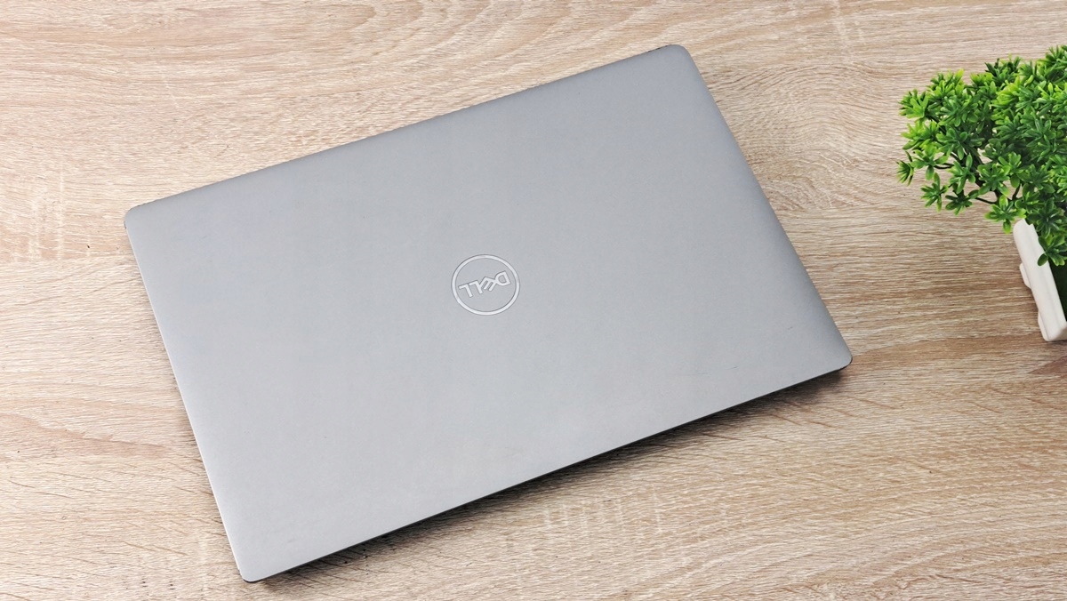 Zamknięty laptop Dell z logo na pokrywie, srebrna obudowa, ustawiony na drewnianym blacie obok rośliny.