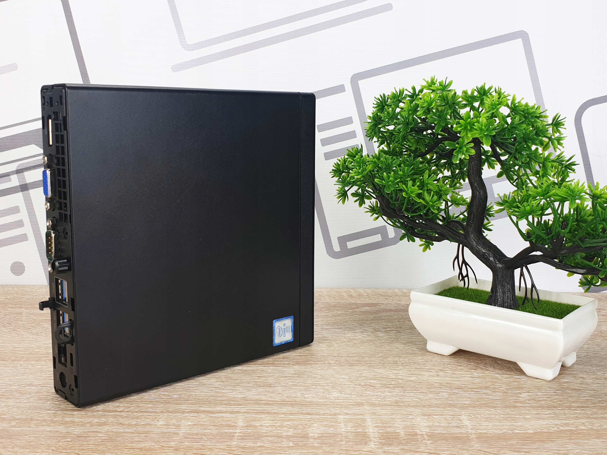 Czarna obudowa komputera HP EliteDesk Tiny stojąca na boku, widoczne porty USB z przodu, obok sztuczne drzewko bonsai.