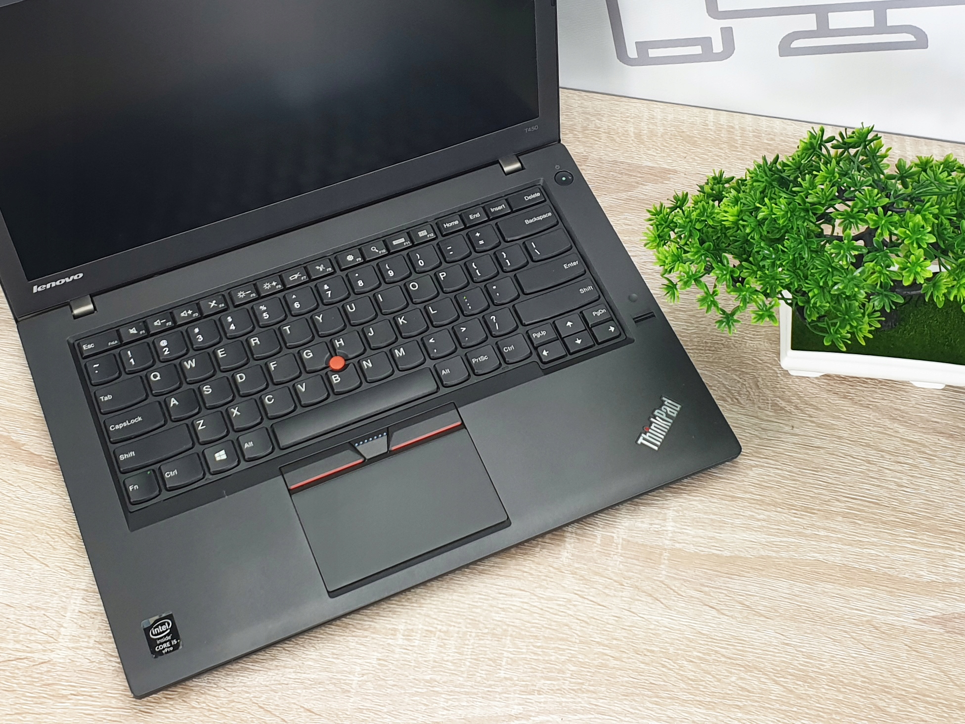 Otwarty laptop Lenovo ThinkPad T450 z widoczną klawiaturą i touchpadem, ekran wyłączony, w tle sztuczna roślina.