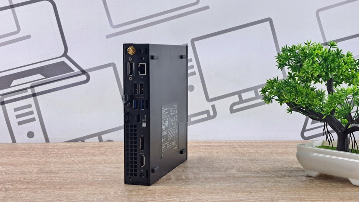 Boczny panel obudowy Dell Micro z widocznymi portami i naklejką, komputer stojący na biurku przy roślinie