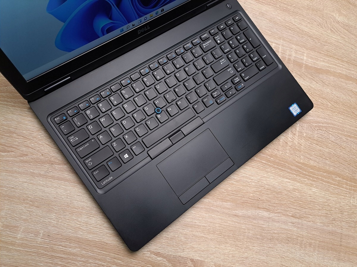 Otwarty laptop Dell Latitude 5580 widziany z góry pod kątem, widoczna klawiatura z touchpadem na drewnianym blacie.