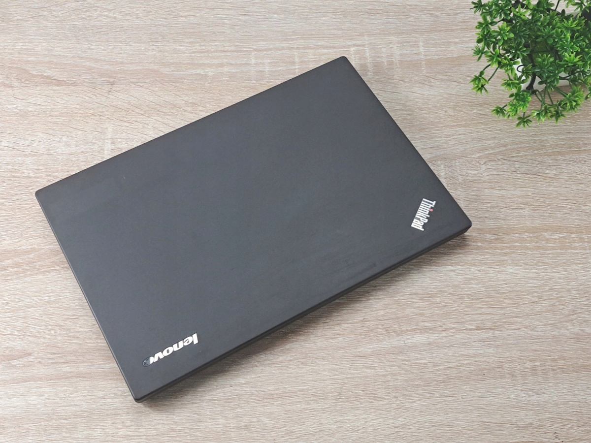 Zamknięty laptop Lenovo ThinkPad z widoczną klapą górną i logo, ustawiony na drewnianym blacie obok rośliny.