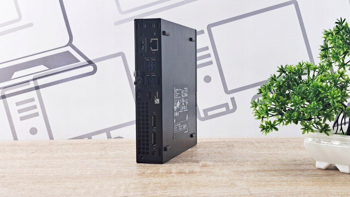 Mini PC Dell 7050 Micro ustawiony bokiem, widoczna tabliczka znamionowa i porty na obudowie, w tle dekoracyjna roślina.