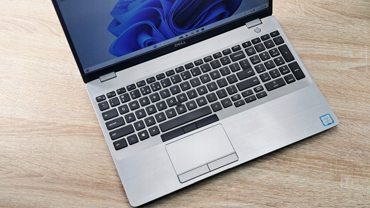 Otwarty laptop Dell Latitude widziany z góry pod kątem, widoczna klawiatura, gładzik i lekko zarysowana obudowa na drewnianym blacie.