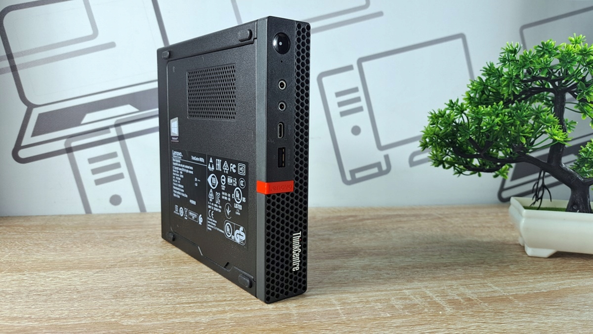 Boczny widok komputera Lenovo ThinkCentre M920Q Tiny z widoczną naklejką z etykietą i czerwonym akcentem na obudowie.