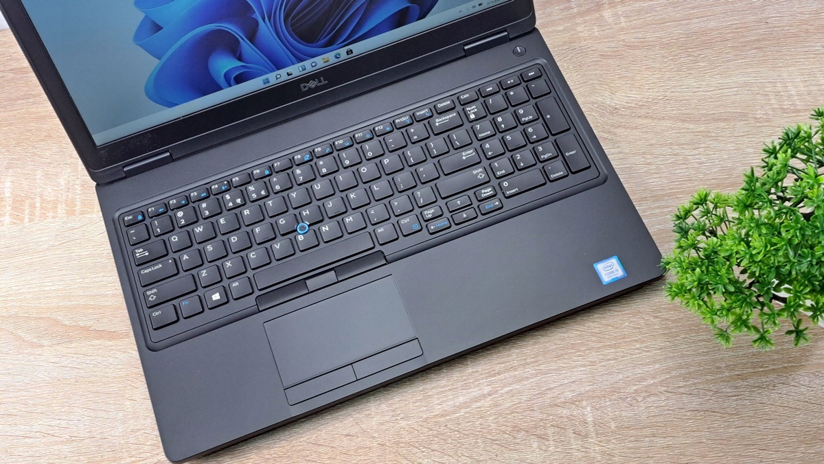 Otwarty laptop Dell Latitude widziany z góry pod kątem, klawiatura i touchpad, obok doniczka z rośliną na drewnianym blacie.