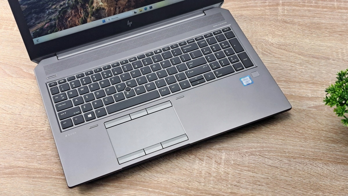 Otwarty laptop HP ZBook widziany z góry pod kątem, widoczna klawiatura z touchpadem i drewniane biurko w tle.