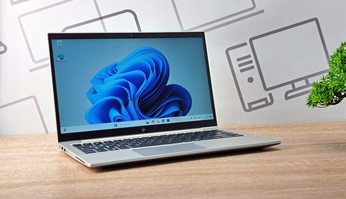Otwarty laptop HP Elitebook ustawiony pod kątem na biurku, ekran włączony z niebieską grafiką systemu Windows 11.