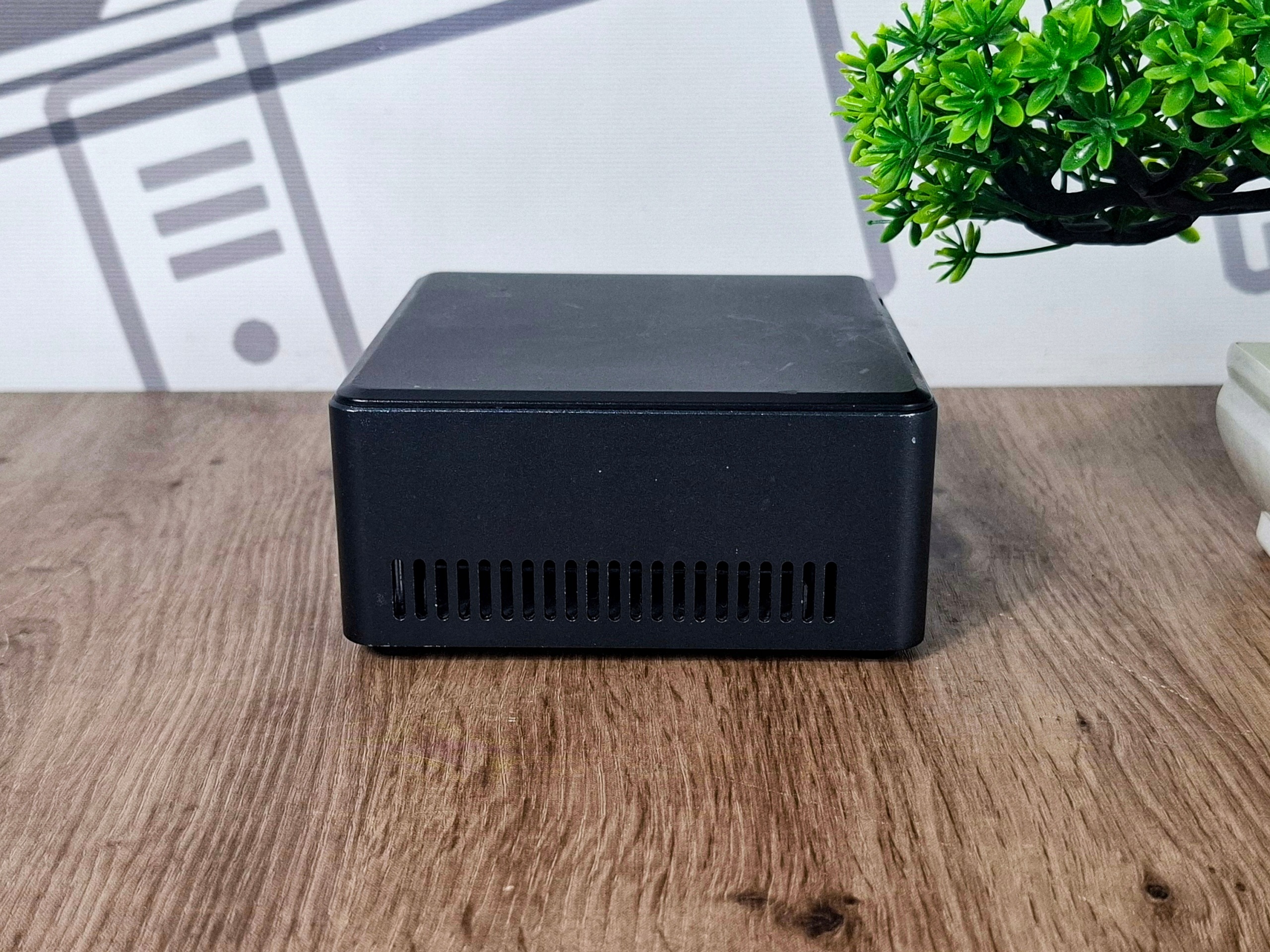 Boczna ściana czarnej obudowy mini PC Intel NUC z otworami wentylacyjnymi, na tle sztucznej rośliny.
