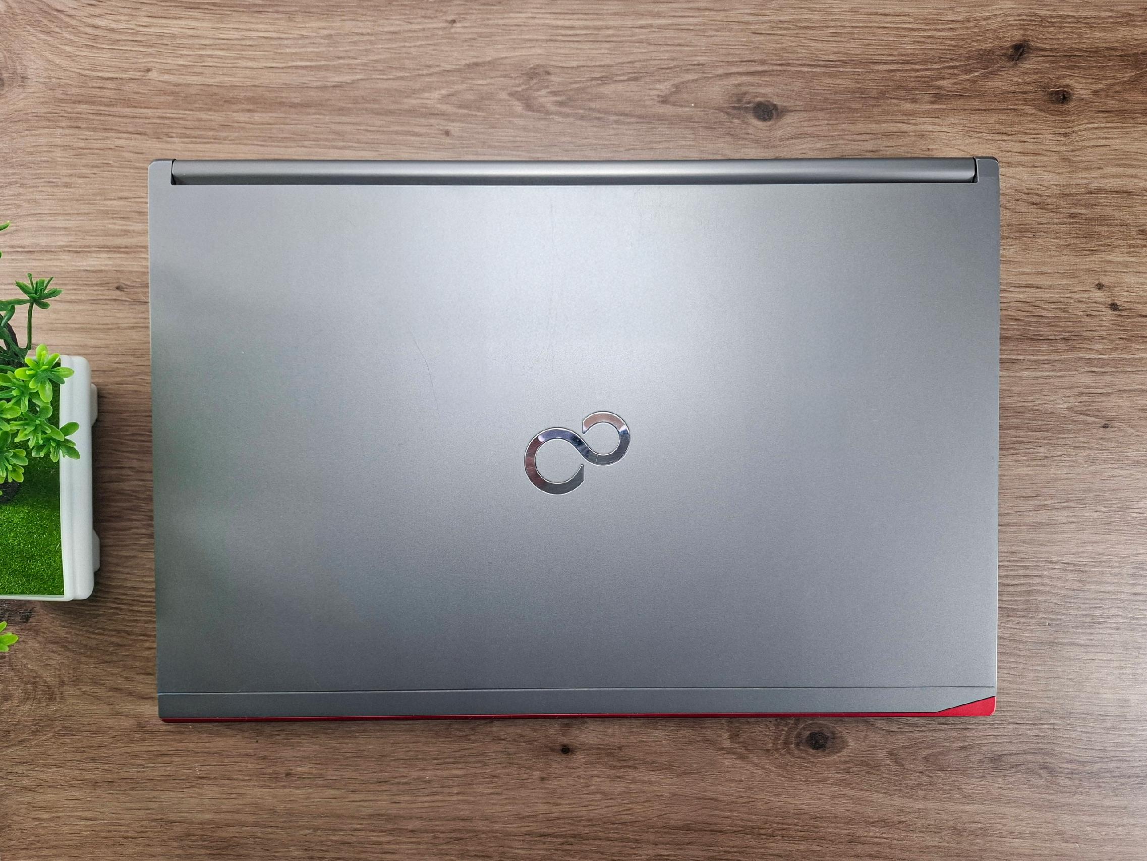 Zamknięta klapa laptopa Fujitsu z logo pośrodku, widok z góry na drewnianym blacie, obok doniczka z rośliną.