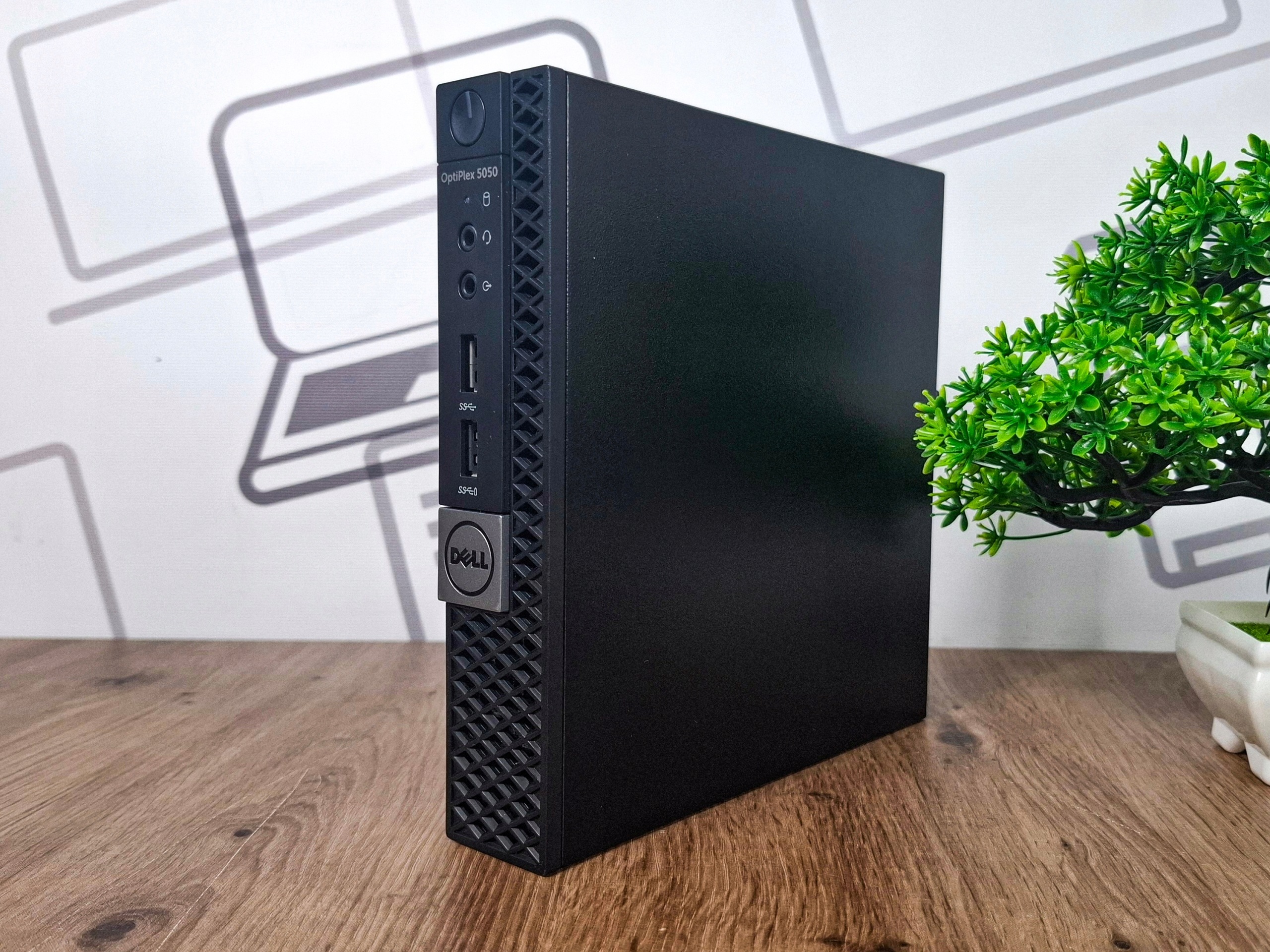 Komputer Dell OptiPlex Tiny stojący pionowo, widok z przodu na perforowaną obudowę i przyciski portów.