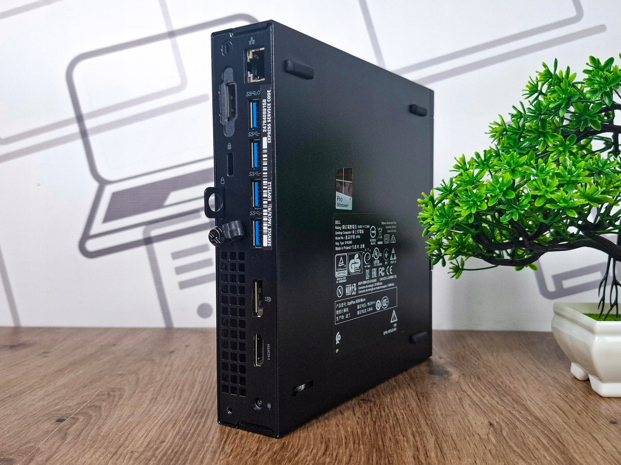 Boczno-tylny widok mini komputera Dell Optiplex Tiny z widoczną tabliczką znamionową i portami, obudowa bez widocznych uszkodzeń.