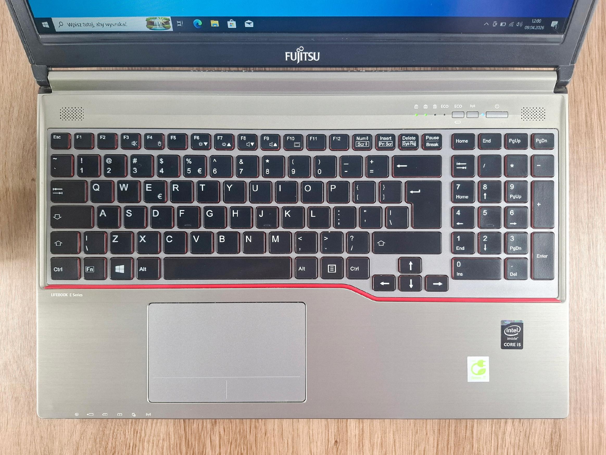 Laptop Fujitsu Lifebook widziany z góry, otwarta klawiatura i touchpad, ekran włączony w tle, obudowa z widocznymi śladami użytkowania.