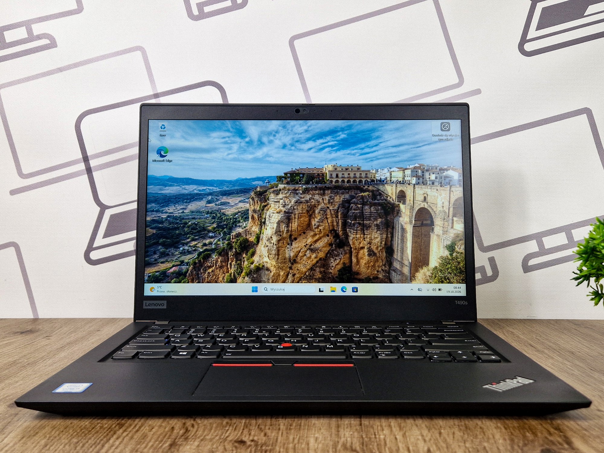 Otwarty laptop ThinkPad T490s na biurku, ekran włączony z widokiem krajobrazu, widoczna klawiatura z czerwonym trackpointem.